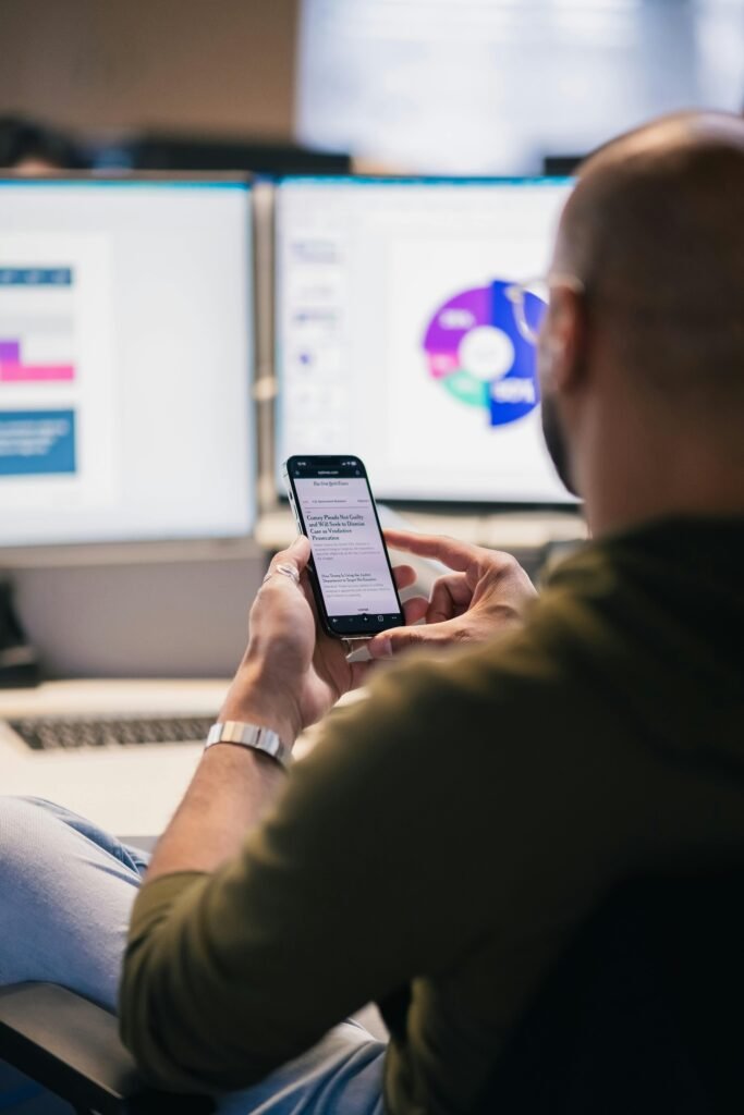 A business professional uses a smartphone while analyzing data on computer screens in a modern office.